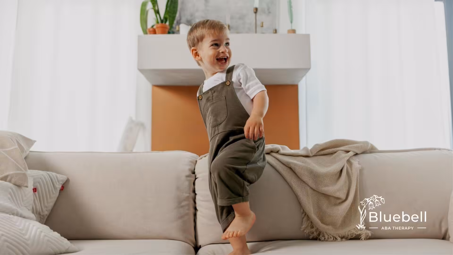 A young child with autism standing playfully on a couch in a bright living room.