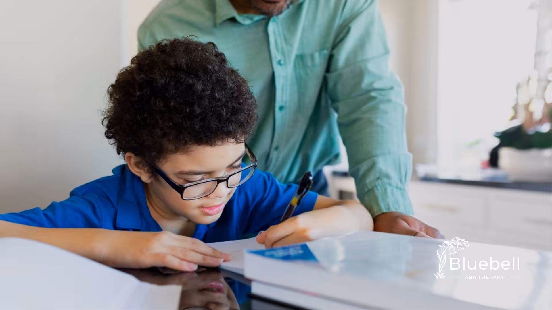 BCBA helping a child with glasses write at a table during ABA therapy.